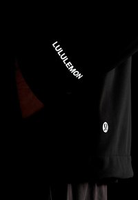 Juodas prailgintas sportinis marškinėliai su balta "LULULEMON" užrašu ant rankovės ir logotipu kairiajame apatiniame kampe. Lygus audinys, prigludusi siluetas.
