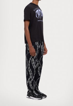 Black cotton t-shirt with white graphic design, paired with black sweatpants featuring chain pattern and text, worn with black sneakers.