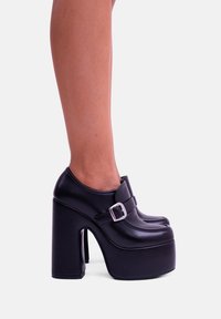 Scarpe con plateau nere, con punta arrotondata, una texture simile alla pelle liscia e un dettaglio con fibbia argentata sul davanti. Design con tacco alto a blocco.