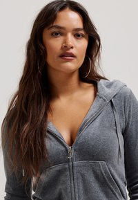 Grau zip-up Kapuzenpullover aus weichem Material, ausgestattet mit einem silbernen Reißverschluss, Fronttaschen und einem dezent gestickten Logo.