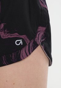 Mustat shortsit, joissa on violetti marmoroitu kuvio ja pieni sivuhalkio. Toisella puolella helman lähellä näkyy pieni valkoinen logo.