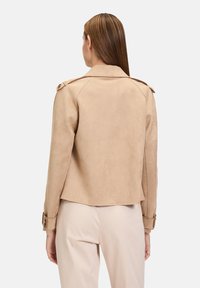 Veste en suédine beige avec un col structuré, des épaulettes et une coupe ajustée, présentant un dos légèrement évasé et un ourlet arrondi.