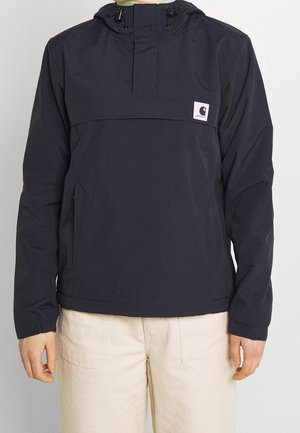 Persona che indossa un anorak Carhartt con cappuccio blu navy e cerniera frontale, con pantaloni beige, in piedi davanti a uno sfondo chiaro e uniforme.