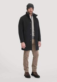 Dunkelgrauer Wollmantel mit hohem Kragen, fünf Knöpfen und Seitentaschen, getragen über einem gemusterten Pullover, kombiniert mit beigen Cordhosen und braunen Stiefeln.