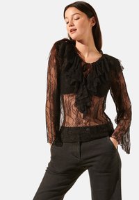 Blouse en dentelle noire avec des accents froncés, tissu transparent, manches longues et un col enkeyhole avec un nœud. Assortie à un pantalon sombre.