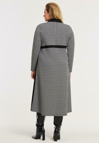 Eine Frau trägt einen langen Mantel mit schwarz-weißer Hahnentritt-Musterung und schwarze Stiefel. Sie steht mit dem Rücken zu einer schlichten Hintergrund.