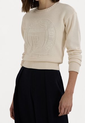 Femme portant un pull en maille beige avec un design en relief de blason et des poignets côtelés, associé à un pantalon noir taille haute.
