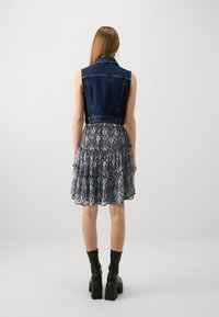 Jeansväst med en figurnära design över en mönstrad, lager-på-lager kjol i mörka färger. Kjolen har volanger och en flödande struktur. Svarta klackstövlar.