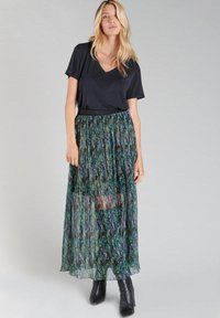 Haut noir à col en V associé à une jupe longue plissée verte avec des motifs multicolores. L'ensemble est complété par des bottines noires.