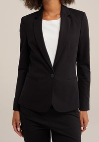 Kvinna klädd i en skräddarsydd svart blazer över vit topp och svarta byxor, stående mot en enkel beige bakgrund.