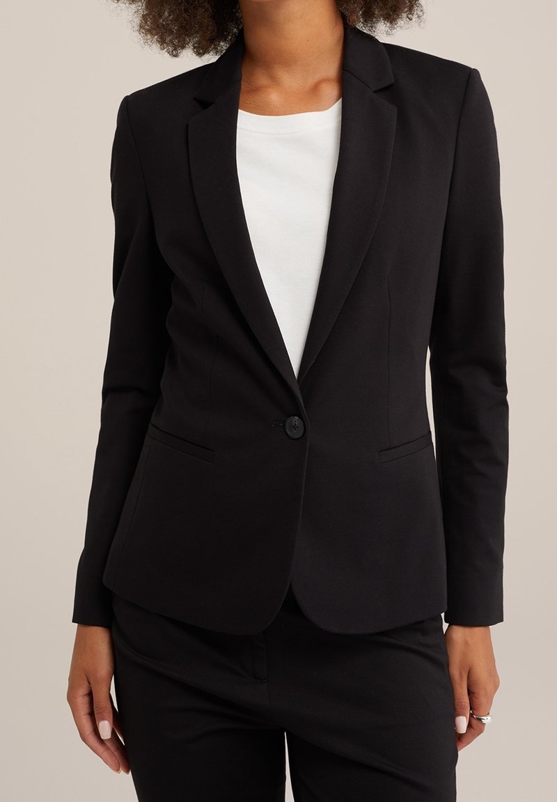 Kvinna klädd i en skräddarsydd svart blazer över vit topp och svarta byxor, stående mot en enkel beige bakgrund.