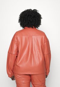 Giacca in similpelle color ruggine con una vestibilità comoda, caratterizzata da bottoni a pressione e un design a pannelli. Superficie testurizzata e dettagli a piega sono visibili.