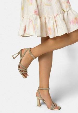 Vrouw draagt gouden sandalen met hoge hakken en enkelbandjes, gecombineerd met een lichte, gerimpelde rok tot op de knie met pastelkleurige patronen.