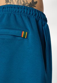 Calças de moletão azul em algodão com um cós elástico, apresentando um pequeno bolso com detalhes em tiras vermelhas, amarelas e verdes.