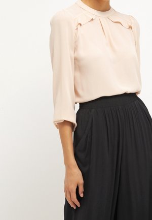 Kobieta w bladoróżowej bluzce z długim rękawem i plisami oraz luźnych, czarnych spodniach z wysokim stanem, stojąca na jednolitym białym tle.