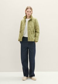 Gepolsterte, olivgrüne Jacke mit hohem Kragen, Frontreißverschluss und Druckknöpfen, kombiniert mit dunkelblauen weiten Jeans und beigen klobigen Schuhen.
