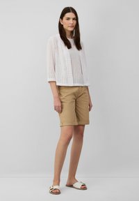 Witte gebreide top met horizontale strepen, gedragen met khaki shorts en beige sandalen met een gouden accent. Model staat tegen een lichte achtergrond.
