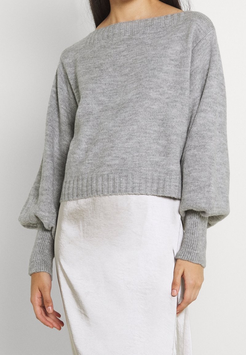 Femme portant un pull en tricot gris clair à manches longues avec des poignets côtelés et une jupe blanche, montrée des épaules aux hanches.