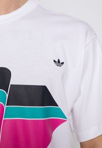 Bílá bavlněná tričko s velkým grafickým designem v černé, růžové a tyrkysové barvě, s malým černým logem Adidas na hrudi.