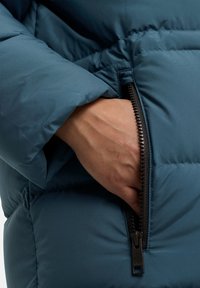 Giacca puffer turchese con tasca con zip. Il tessuto è morbido e leggermente lucido, con un primo piano di una mano che riposa all'interno della tasca.