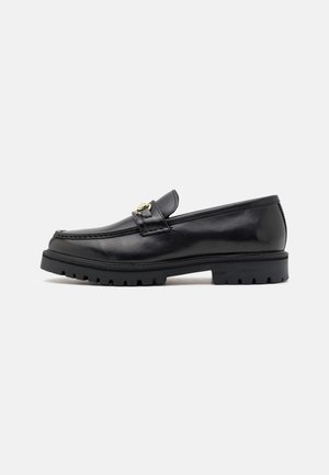 Mocassin en cuir noir avec semelle épaisse en caoutchouc robuste et détail en métal doré sur le dessus, vu de côté sur fond blanc.