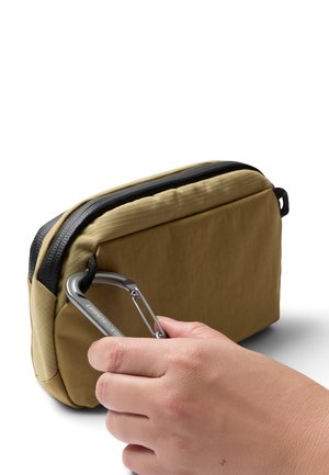 Hand hält einen silbernen Karabiner, der an der Schlaufe eines kleinen, hellbraunen Täschchens mit Reißverschluss vor weißem Hintergrund eingehakt ist.