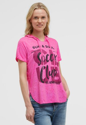 Woman wearing a bright pink short-sleeve hooded t-shirt with black text, paired with blue jeans, standing with one hand in pocket.