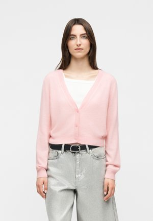 Frau trägt eine hellrosa, geknöpfte Strickjacke über einem weißen Oberteil, kombiniert mit hellgrauen, hoch taillierten Hosen und einem schwarzen Gürtel.