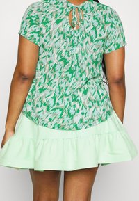 Grønblomstret bluse med korte ærmer og et bånd bagpå, parret med en lys grøn, volangskørt. Glat stof, afslappet pasform.