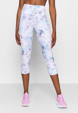 Kobieta nosząca wysokowysokie, skrócone legginsy tie-dye w odcieniach niebieskiego, fioletowego i białego, w połączeniu z różowymi sportowymi butami, stojąca na jednolitym tle.