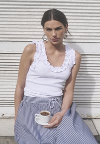 Top blanco sin mangas con cuello con volantes, combinado con una falda a rayas azul y blanca. La modelo sostiene una pequeña taza de café sobre un platillo.