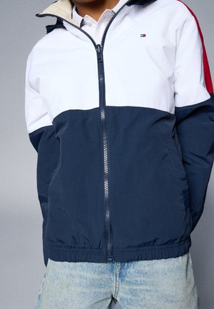 Persona con chaqueta de colores con cremallera en blanco, azul marino y rojo, con vaqueros azul claro, de pie frente a un fondo gris.