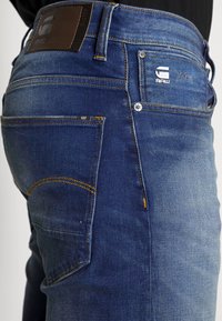 Denimjeans i en medelblå tvätt, med kontrasterande gul sömnad, femfickorsdesign och en brun läderetikett vid midjan.