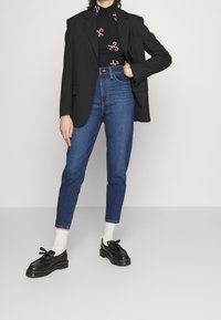 Black blazer over a black shirt with candy cane print, paired with blue skinny jeans and black lace-up shoes, featuring white socks.