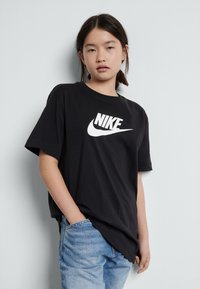 Fiatal ember fekete Nike pólót és világoskék farmernadrágot visel, és egy sima világosszürke falnak támaszkodik, egyik keze a zsebében van.