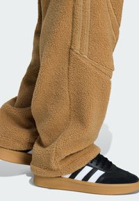 Person in lockeren beigefarbenen Fleecehosen und schwarzen Sneakers mit weißen Streifen und Gummisohlen, stehend vor einem hellen Hintergrund.