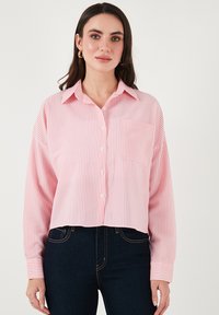Cropped button-up shirt in light pink with white stripes. Features a collar, long sleeves, and a single chest pocket. Fabric appears smooth.