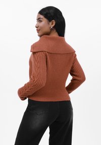 Rustfarget ribbestrikket genser med bred krage og kabelstrikkede ermer, sammen med svarte tettsittende jeans, vist fra en sidevinkel.