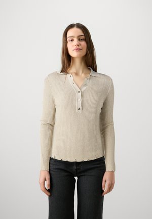Beige geribbelde lange mouwen shirt met een gekraagde halslijn, knoopsluiting en subtiele stiksels langs de zoom. Gecombineerd met donkerblauwe jeans.