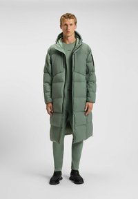 Langer grüner Daunenmantel mit Kapuze, ausgestattet mit einem Reißverschluss und gestepptem Design. Getragen über einem passenden Outfit und schwarzen Sneakers.