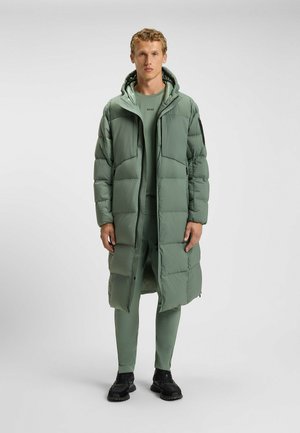 Lång grön puffjacka med huva, med dragkedjestängning och quiltat mönster. Bärs över en matchande outfit och svarta sneakers.