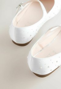 Weiße Ballerinas mit glatter Textur, verziert mit kleinen runden Perlen, ausgestattet mit einem niedrigen braunen Absatz und einem Reißverschluss auf der Rückseite.