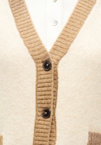 Κοντινό πλάνο κρεμ ζακέτας με καφέ ριμπ τελείωμα και μεγάλα σκούρα κουμπιά πάνω από λευκό ριμπ πουκάμισο.