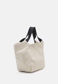 Borsa tote in tela beige con ampi manici neri, caratterizzata da un marchio in rilievo, in piedi su uno sfondo bianco uniforme.