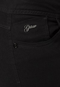 Jeans de mezclilla negra con un bolsillo delantero, que presentan un logotipo plateado de G-Star. Textura suave y costura estándar visible.