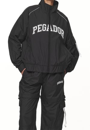 Persona con un chándal negro Pegador con chaqueta con cremallera y pantalones cargo, con las manos en los bolsillos, de pie frente a un fondo blanco.