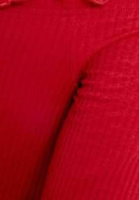 Červená žebrovaná látka s měkkou texturou, charakterizovaná přiléhavým střihem a viditelnými horizontálními pruhy. Bez kovových doplňků či ozdob.