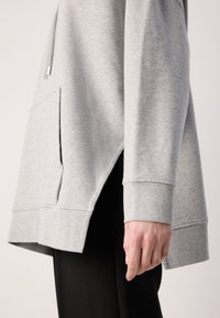 Hanorac gri cu buzunar pe lateral, manșete ributate și croială lejeră. Materialul pare moale și texturat, completând o pereche de pantaloni negri.