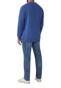 Uomo con felpa blu a maniche lunghe, jeans blu a vestibilità dritta e sneakers bianche, in piedi con le spalle rivolte verso la camera su sfondo bianco.