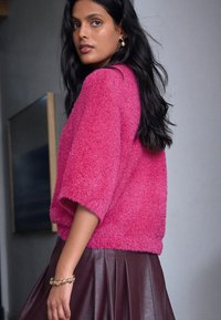 Švelnus, trumpomis rankovėmis rožinis megztinis derinamas su platėjančia tamsiai rudos spalvos sijonu su klostėmis. Ypatingas minkštas audinys ir laisvas siluetas. Aksesuaras – auksinė apyrankė.